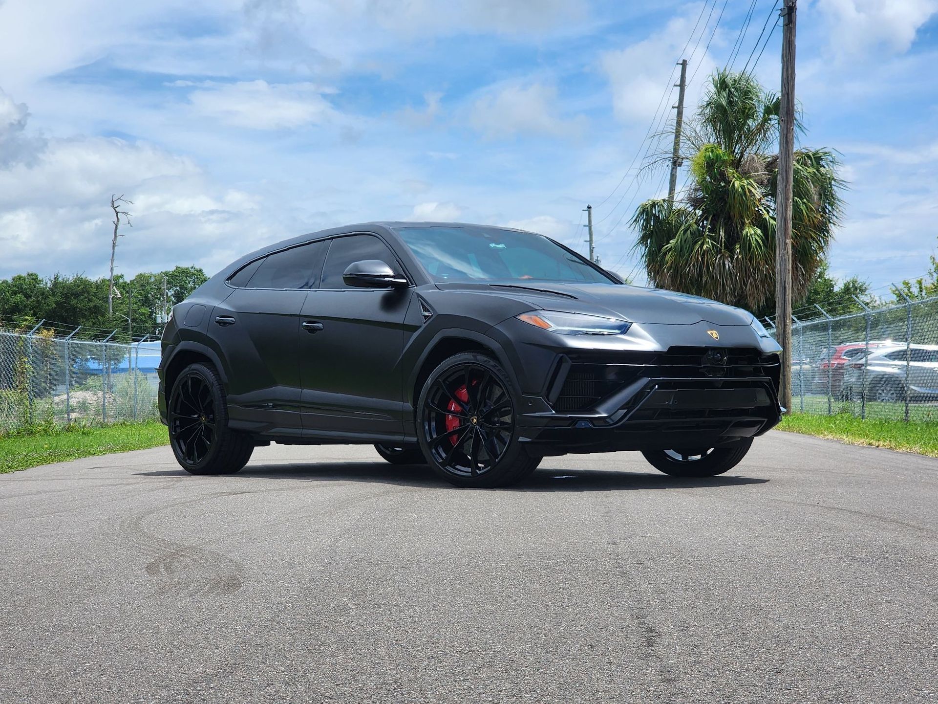 Black Lamborghini Urus SUV parked on a paved road, red brake calipers, black wheels, against a blue sky.