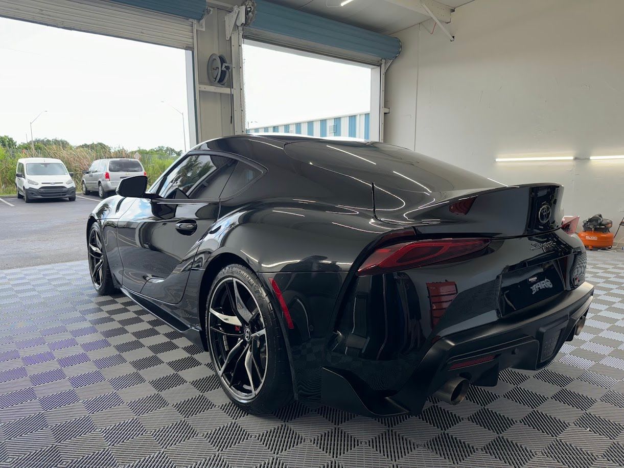 Black Toyota Supra sports car parked on a checkered floor in a bright garage.