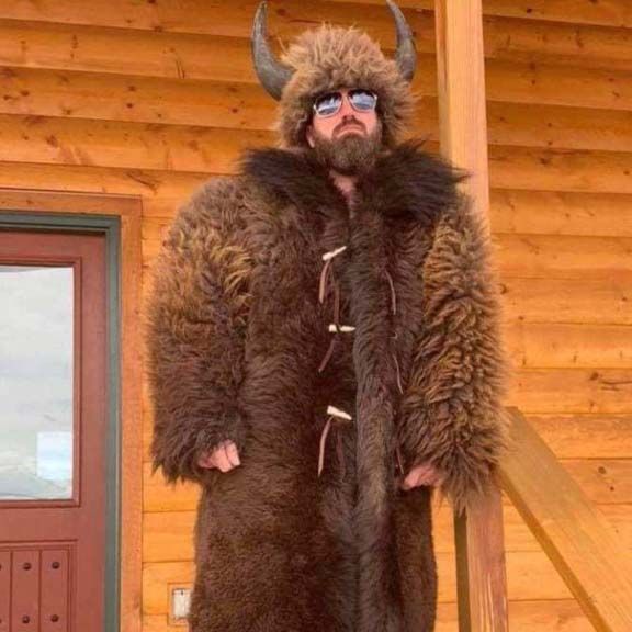 A man wearing a fur coat and horned hat is standing in front of a log cabin.