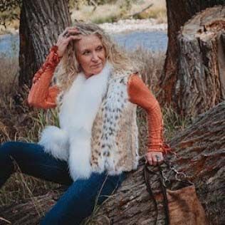 A woman is sitting on a log in the woods wearing a fur vest.
