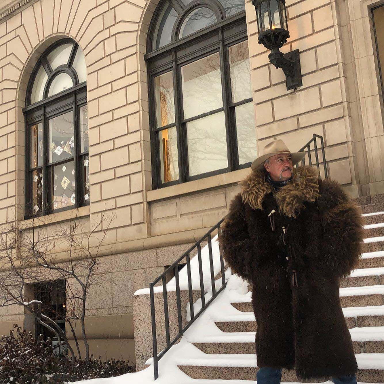 A man in a fur coat and cowboy hat is standing on stairs in front of a building
