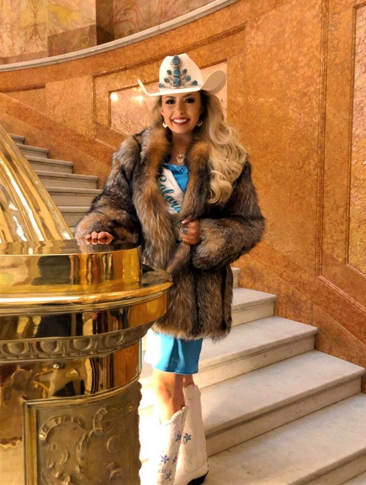 A woman in a fur coat and cowboy hat is standing on a set of stairs.