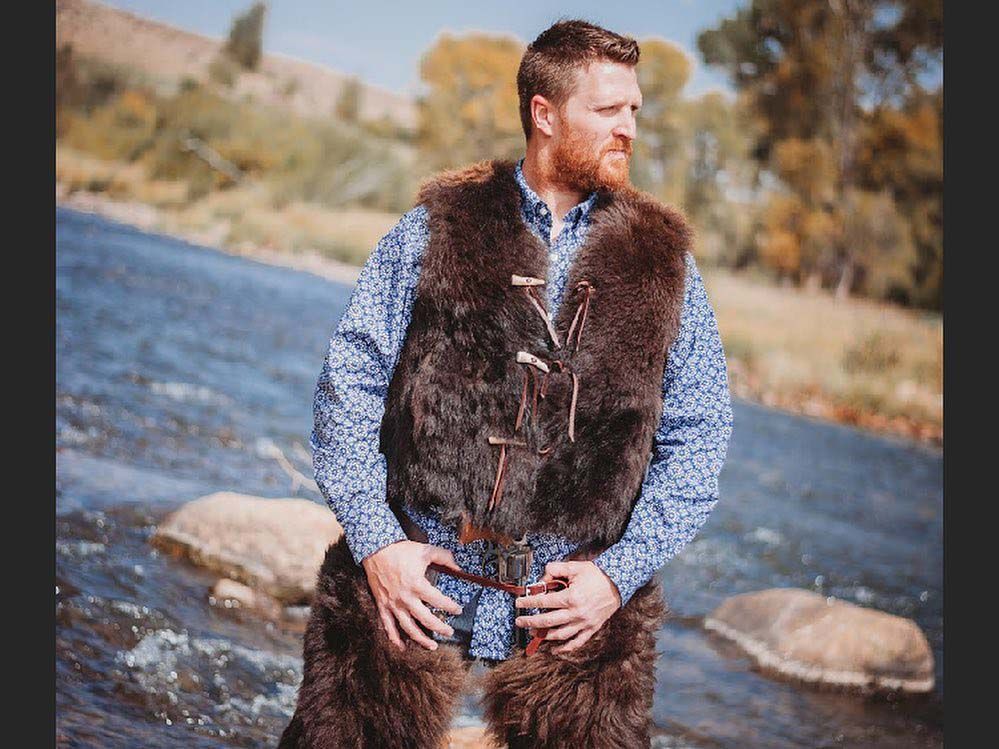 A man wearing a fur vest is standing next to a river.