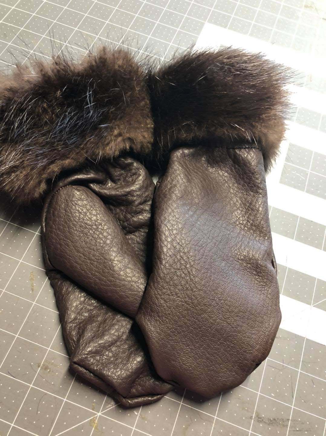 A pair of brown leather mittens with fur trim are sitting on a cutting board.
