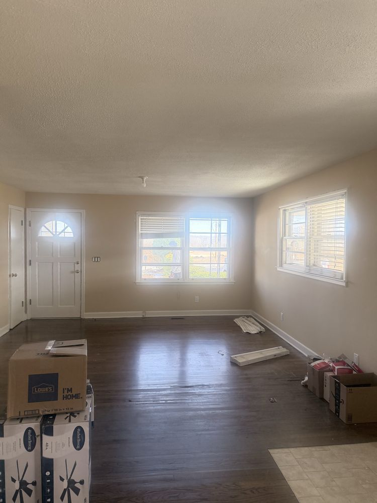 Empty living room with hardwood floors, white walls, windows, and a front door; boxes and trim pieces on the floor.