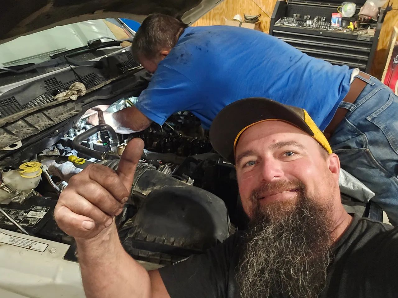 A bearded man gives a thumbs-up while a colleague works on the engine of a car in a garage.