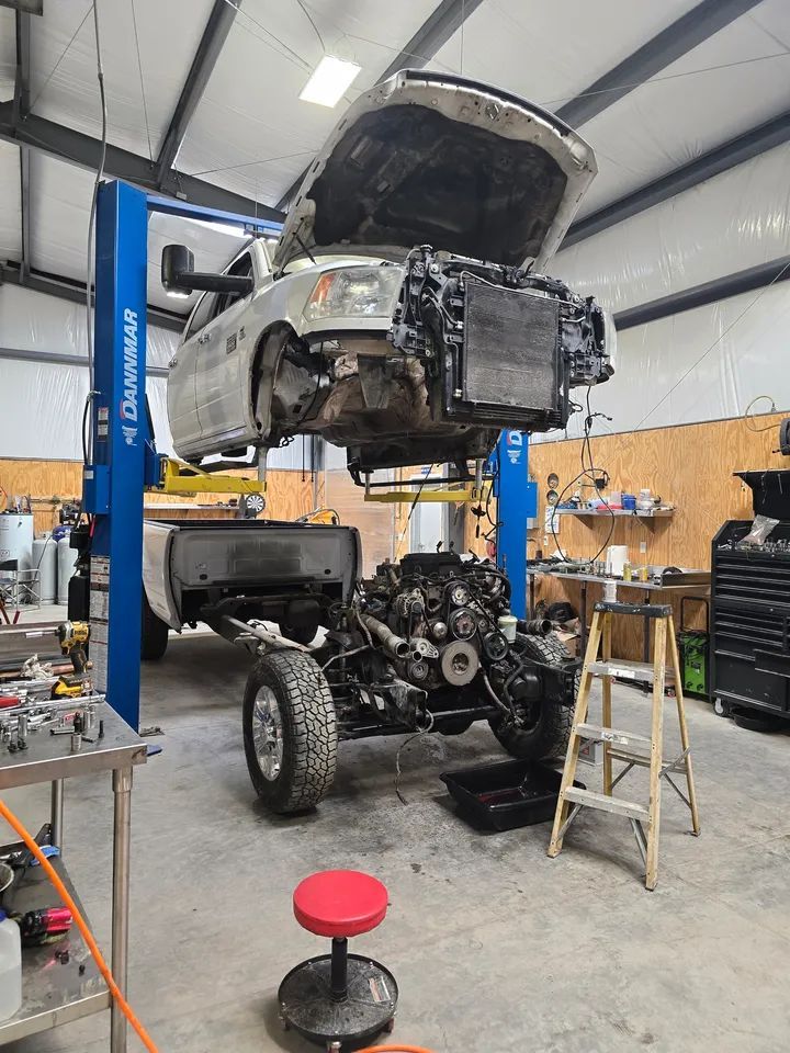 A silver pickup truck body is lifted on a two-post hydraulic lift, separated from its engine and frame in a workshop.
