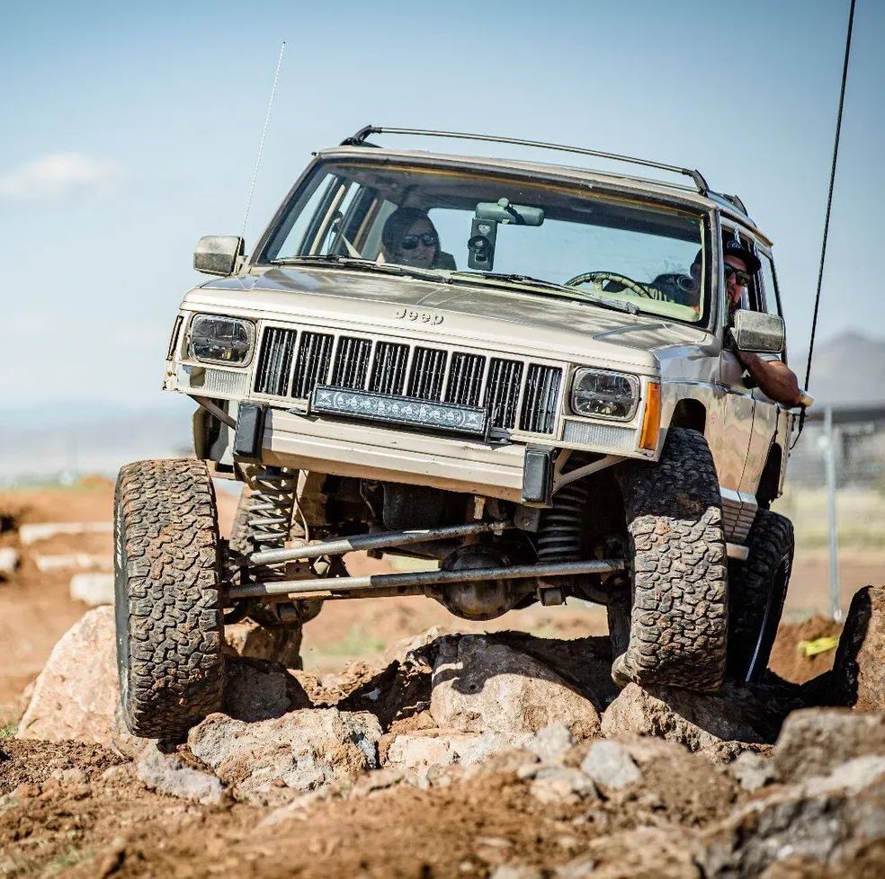 A beige Jeep Cherokee off-roading over rocky, uneven terrain under a clear blue sky.