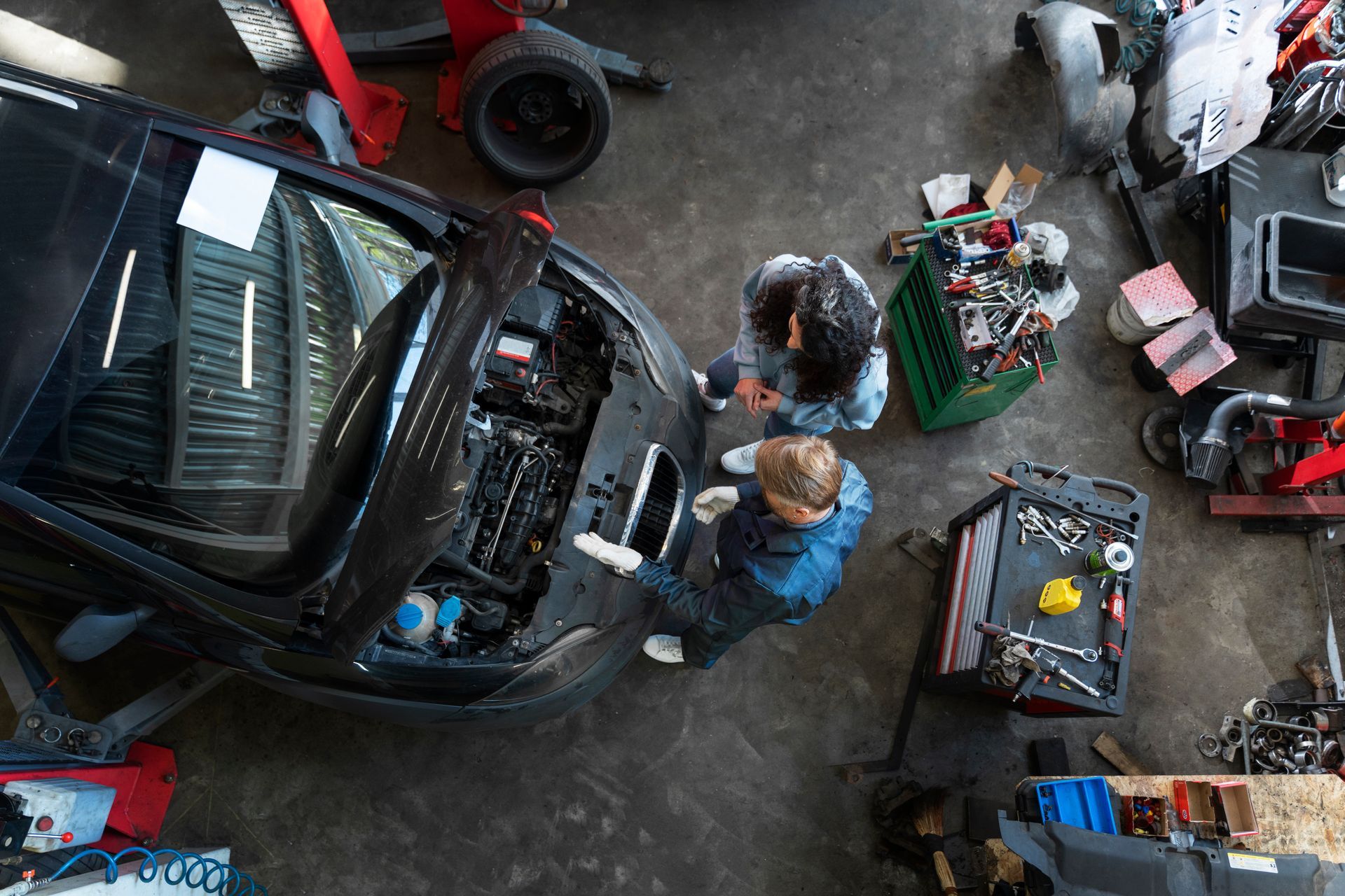 Two mechanics wearing gloves examine the open engine bay of a black car in an organized automotive workshop.