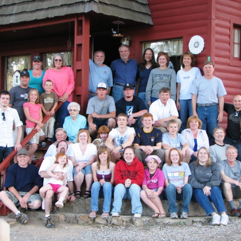 Large group posing on cabin porch and steps, with red wooden walls in the background