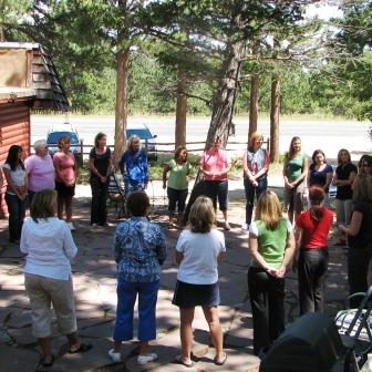 People standing in a circle outdoors by trees, appearing to participate in a group discussion or activity.