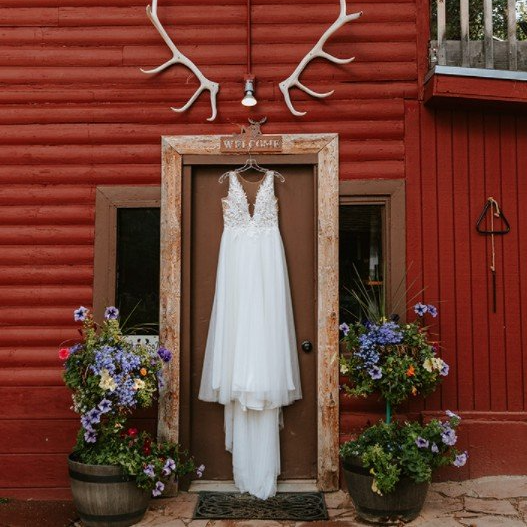 White wedding dress hanging in a rustic wooden frame, flanked by flower pots against a red barn wall