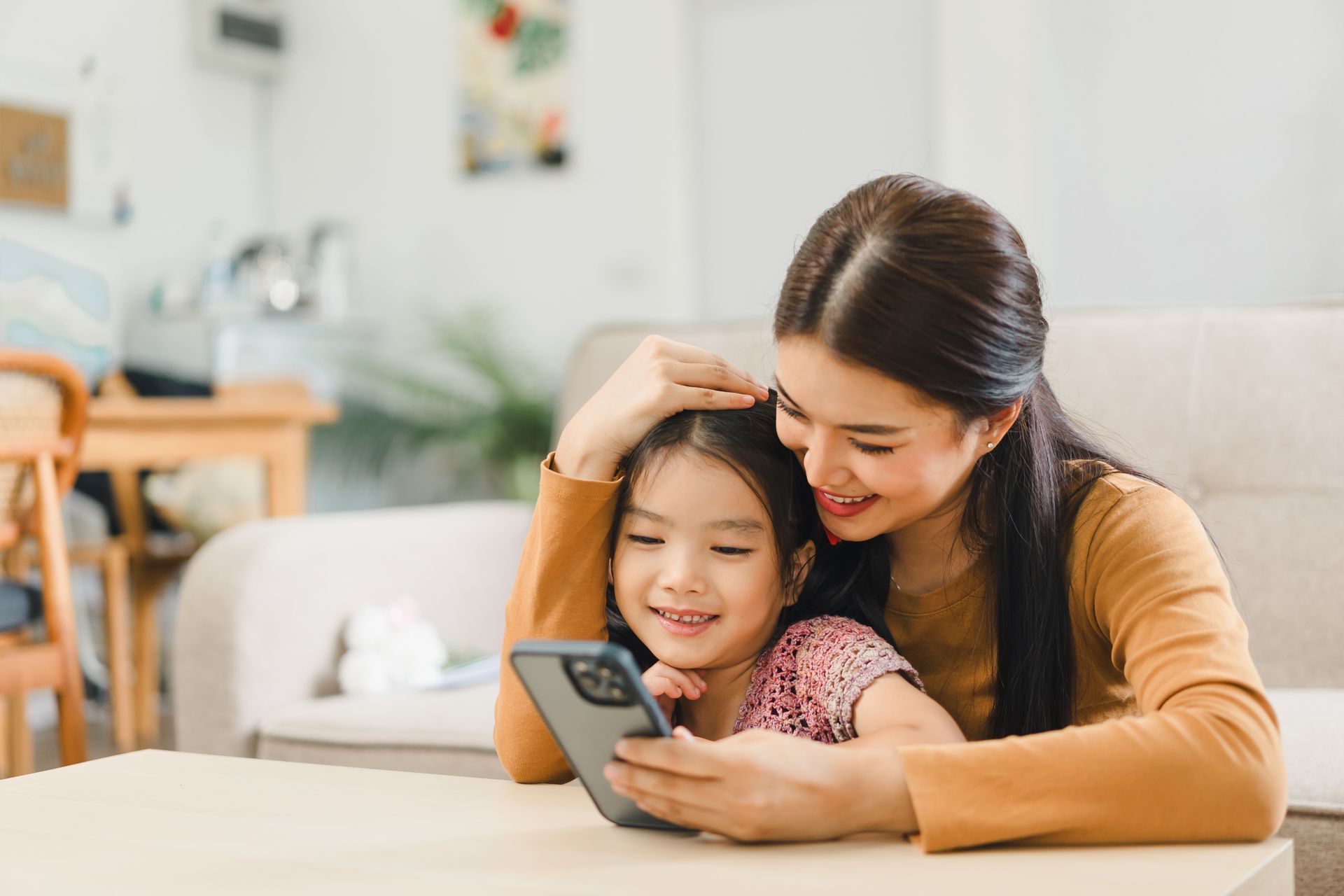 Mom and daughter smiling and looking at a phone. They are seated in a living area.