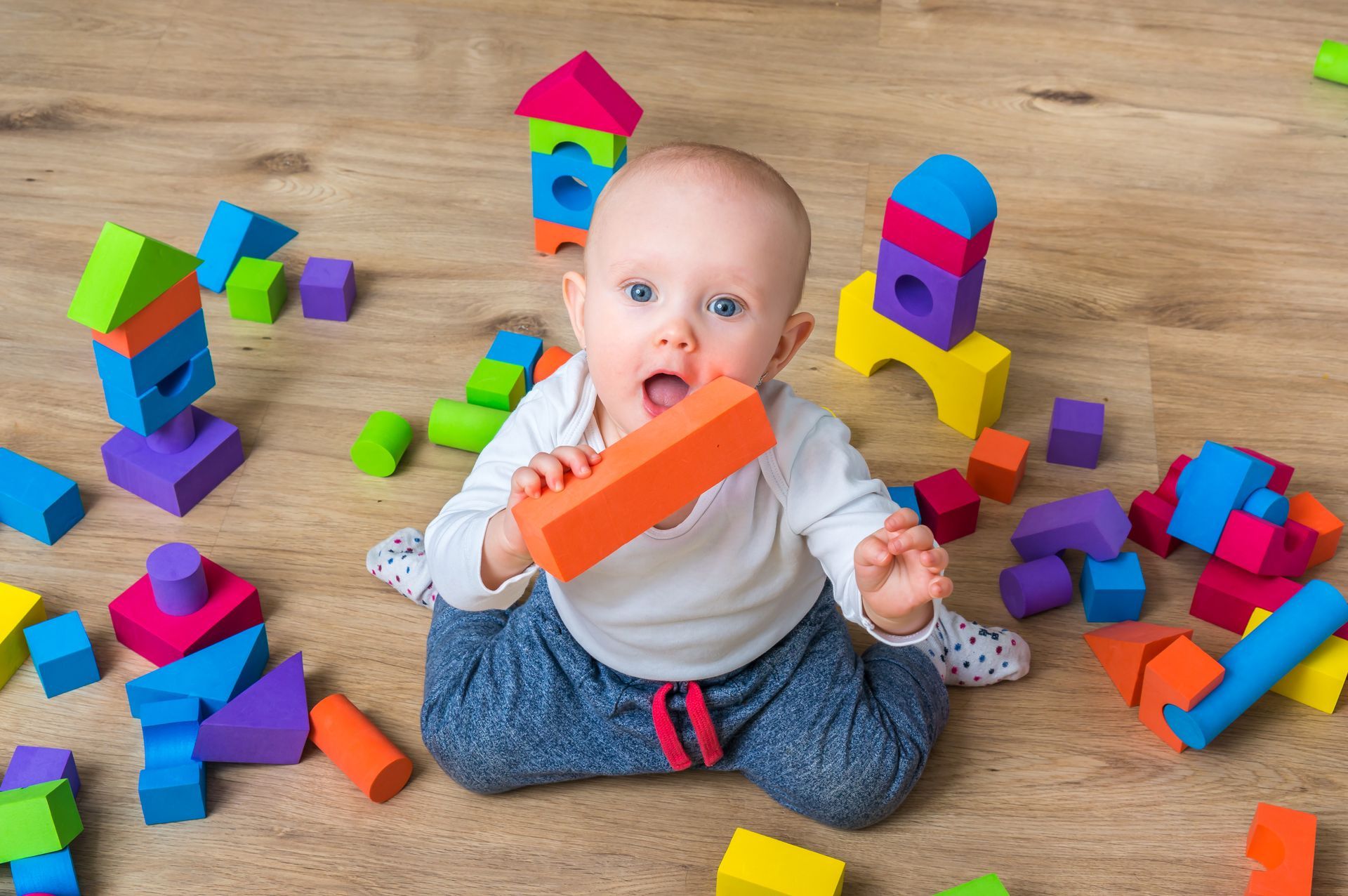 A baby is sitting on the floor looking up at the camera and surrounding by colorful blocks.