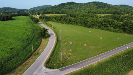 A winding road through green fields and forested hills, with hay bales scattered in the pasture.