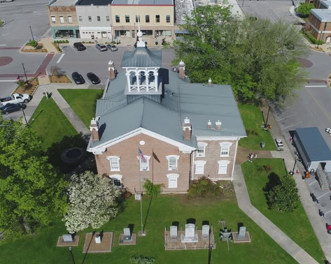 Aerial view of a small brick church with a gray roof and cemetery graves in front, near a street and parking lot.