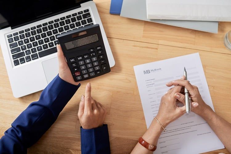 Hands using a calculator and pen over financial paperwork on a desk beside a laptop.