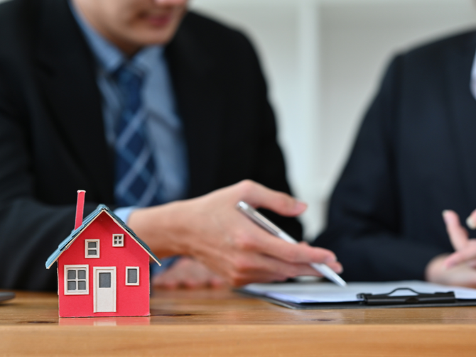 Small red house model on a table with business people in suits discussing in the background