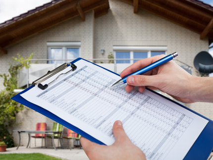 Hand holding a clipboard and pen in front of a house for an inspection checklist