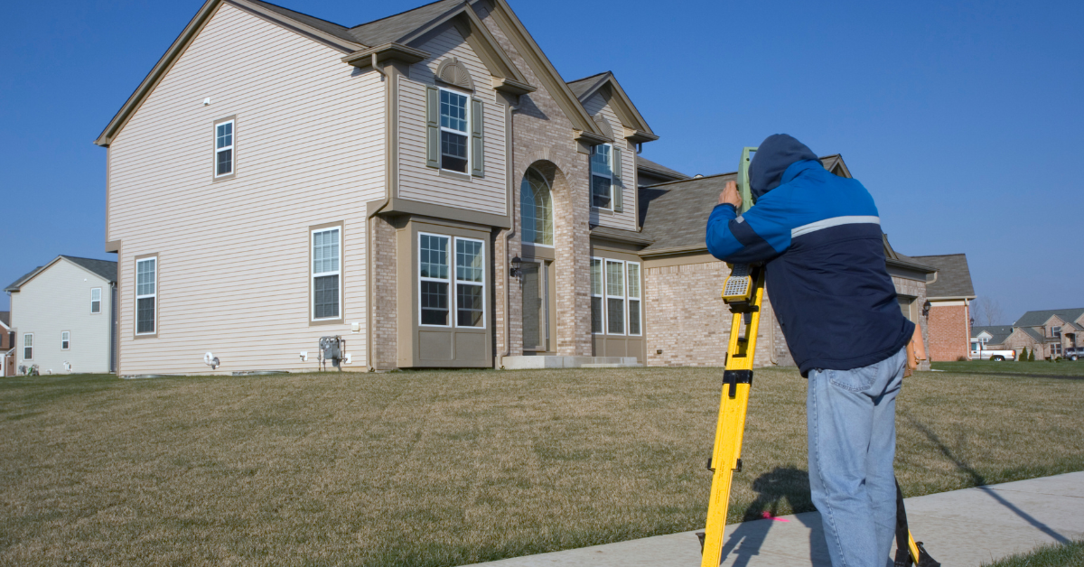 Surveyor using a tripod instrument in front of a beige suburban house on a lawn