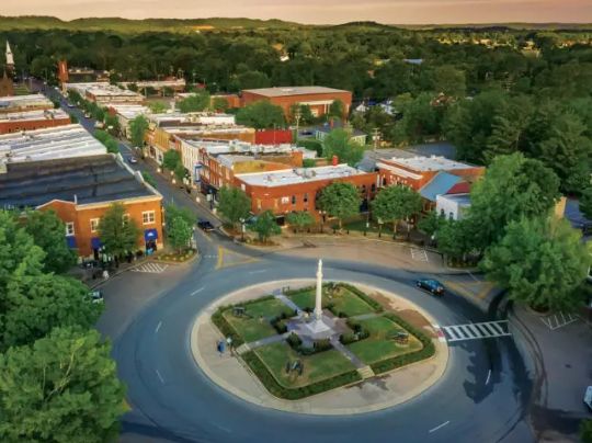 Aerial view of a roundabout with a fountain, surrounded by brick buildings and trees at sunset.