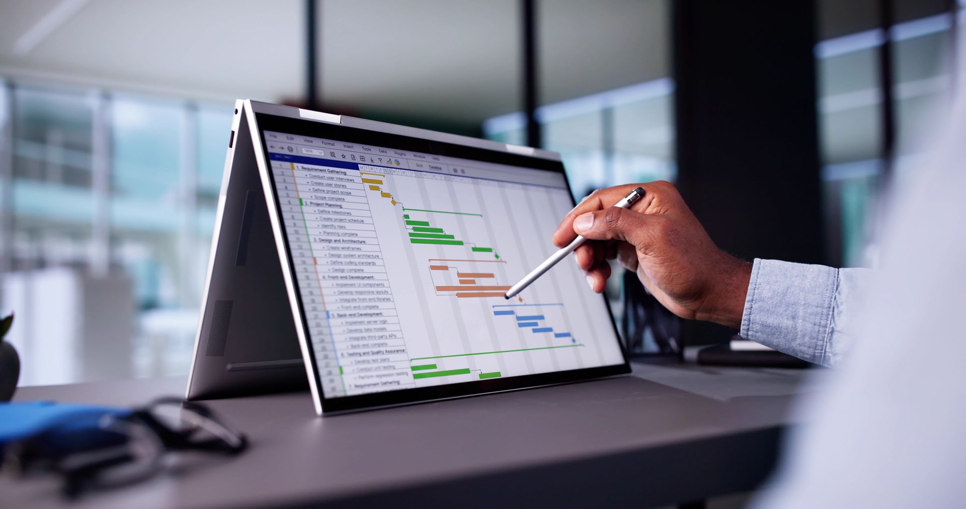 Person uses stylus to interact with a project timeline on a laptop in a modern office. Person uses stylus to interact with a project timeline on a laptop in a modern office.