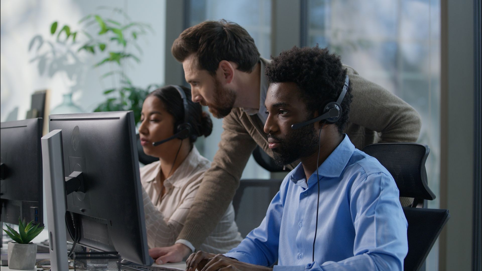 Three people in an office wearing headsets; one man leans over another's shoulder, pointing at a computer screen. Three people in an office wearing headsets; one man leans over another's shoulder, pointing at a computer screen.
