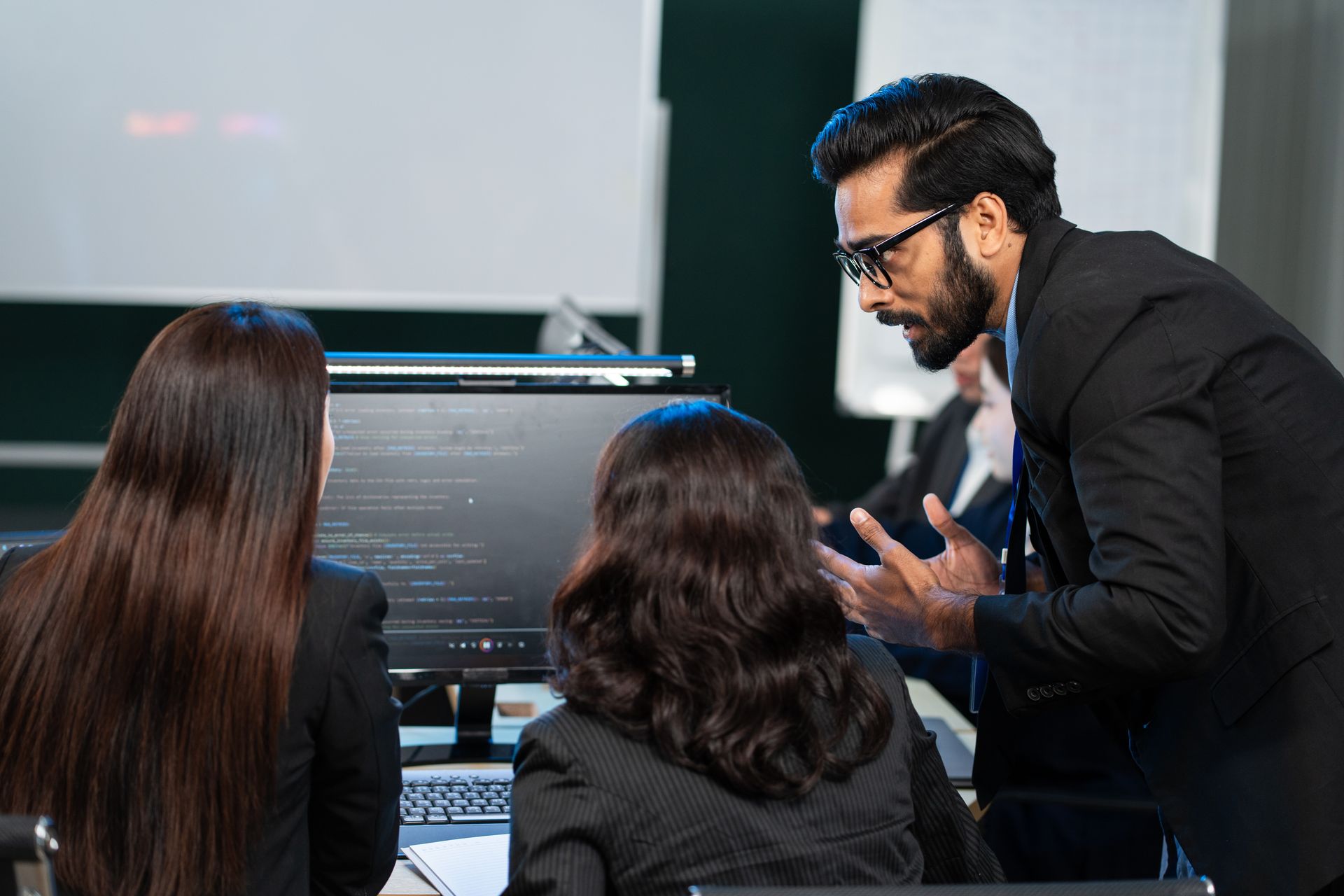 Man in suit gestures at a computer screen as two women look on. A workplace scene. Man in suit gestures at a computer screen as two women look on. A workplace scene.