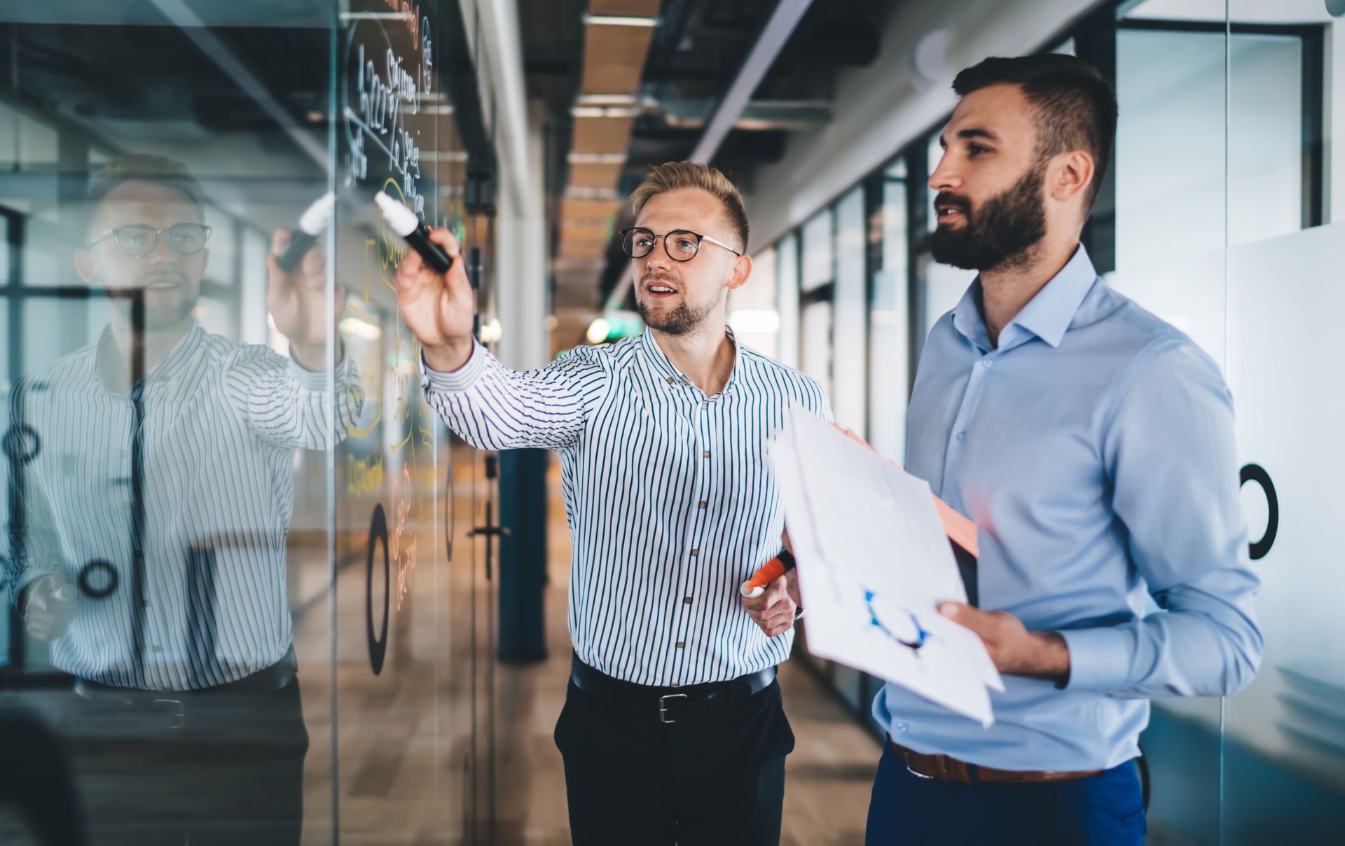 Two men in an office brainstorming on a glass wall; one writes with a marker, the other holds papers. Two men in an office brainstorming on a glass wall; one writes with a marker, the other holds papers.