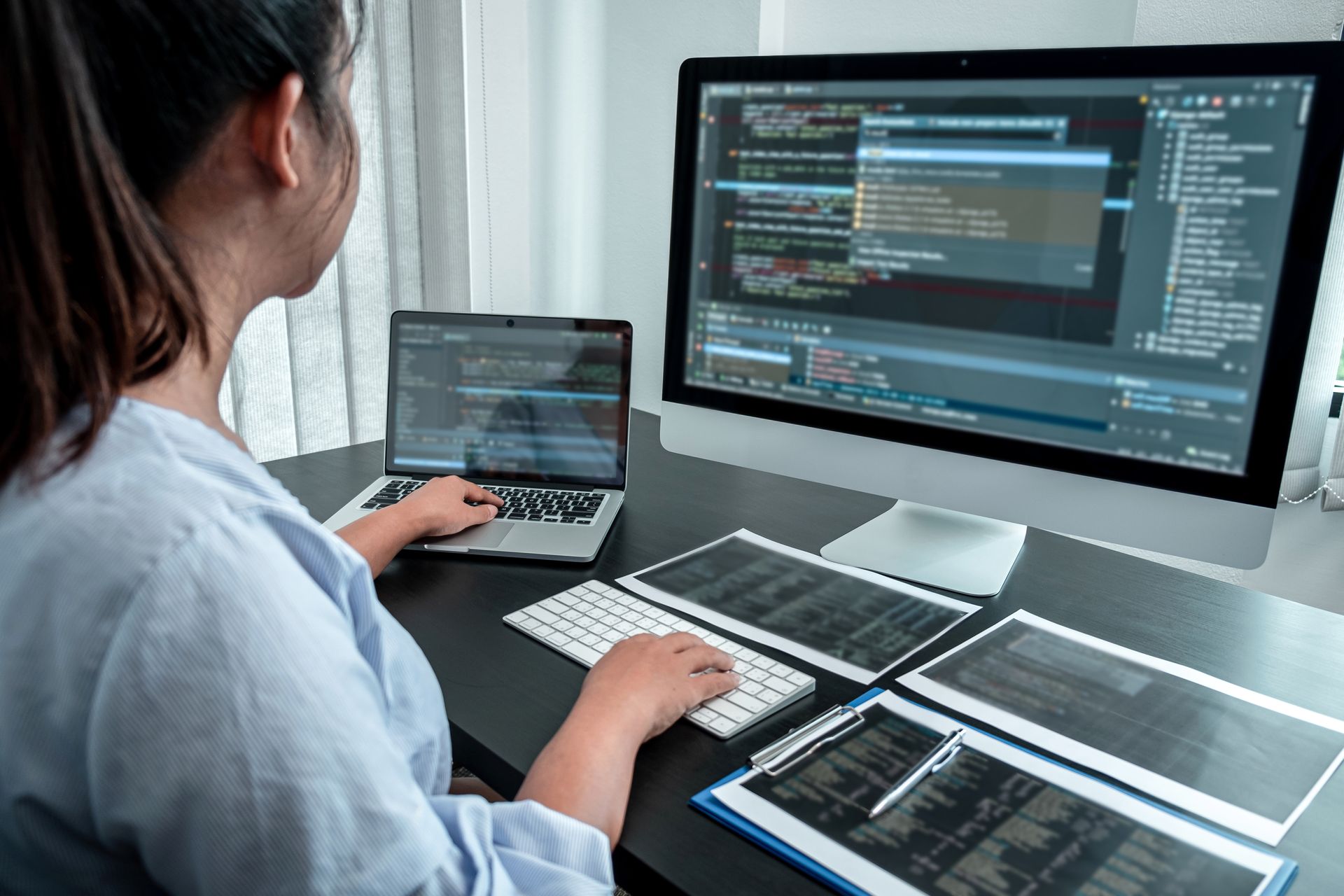 Woman coding on a laptop and desktop, surrounded by code printouts, in a bright office. Woman coding on a laptop and desktop, surrounded by code printouts, in a bright office.
