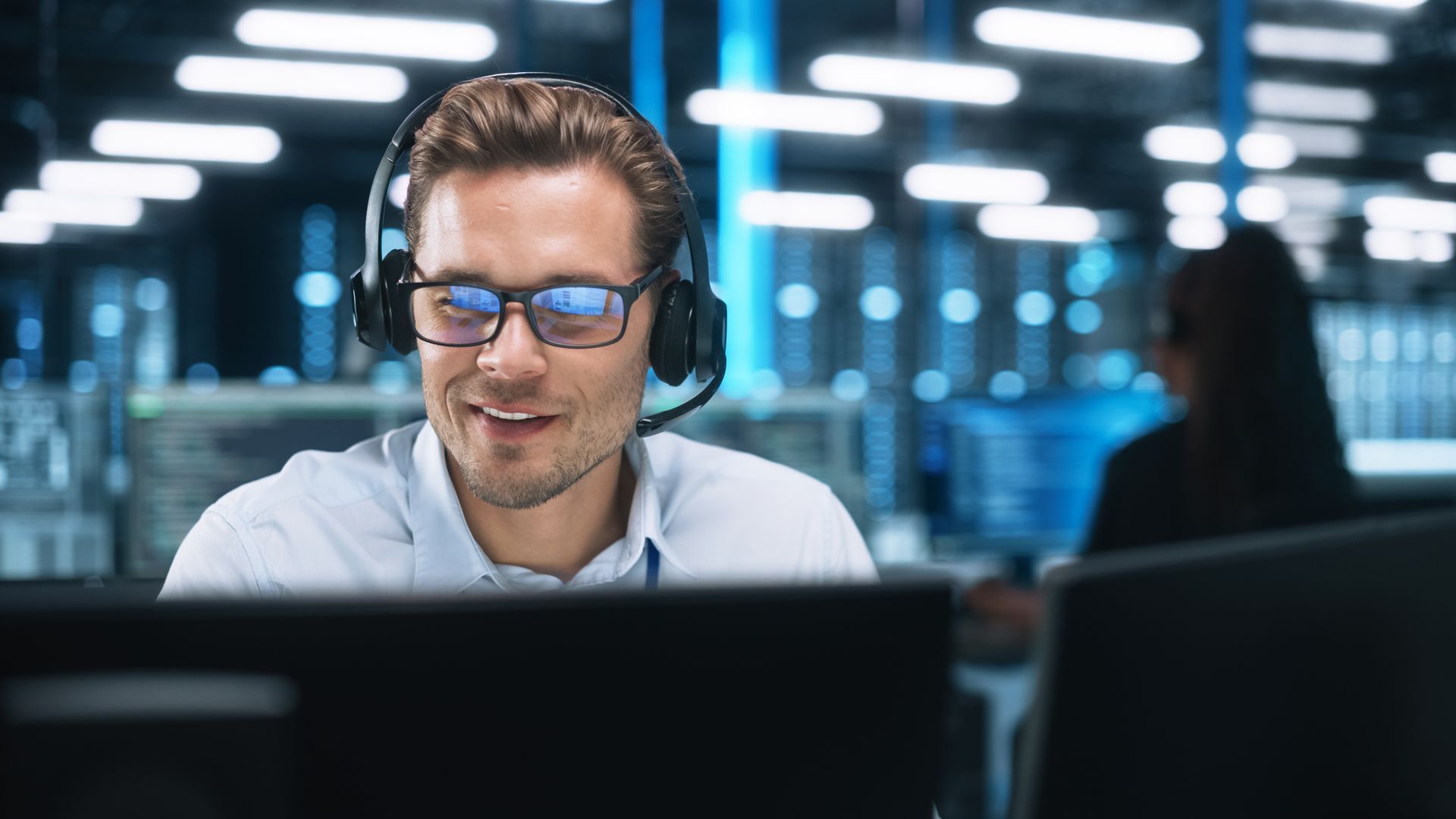 Man with headset and glasses smiling at a computer screen in a server room. Man with headset and glasses smiling at a computer screen in a server room.