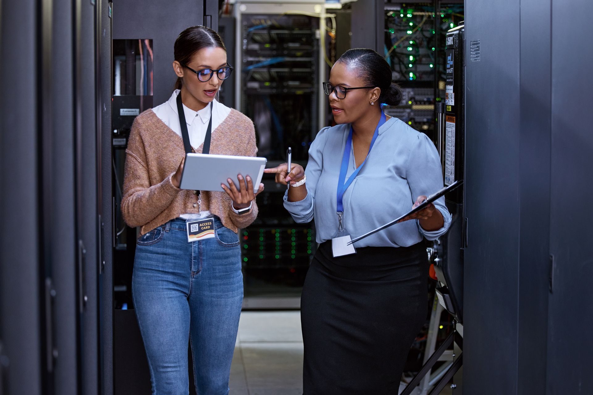 Two women in a server room, one holding a tablet, the other a clipboard, discussing data. Two women in a server room, one holding a tablet, the other a clipboard, discussing data.