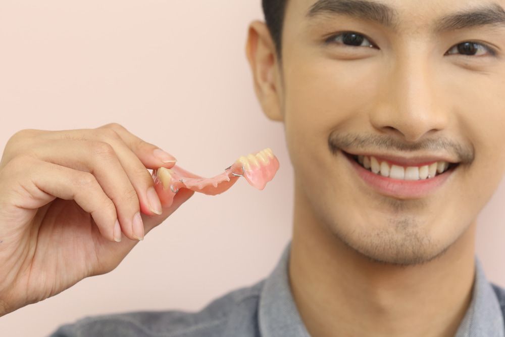 A Man Is Holding A Denture In His Hand And Smiling — Highlands Denture Clinic in Willow Vale, NSW