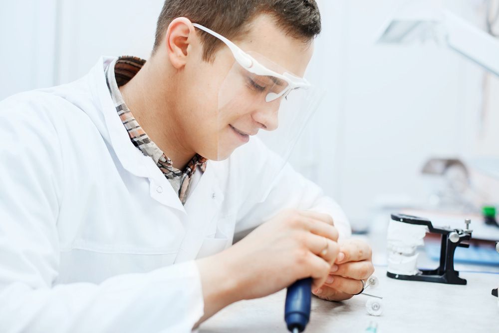 A Man In A Lab Coat And Goggles Is Working On A Piece Of Equipment — Highlands Denture Clinic in Willow Vale, NSW