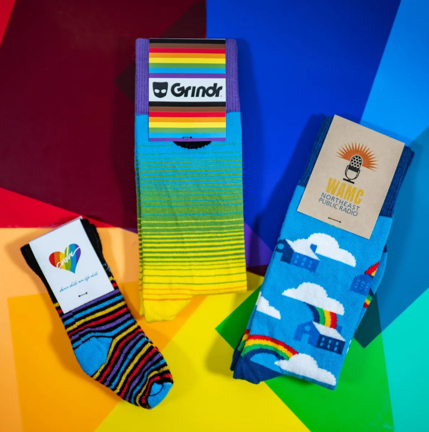 Three pairs of rainbow-themed socks: Grindr socks, house socks with a rainbow, and a heart-shaped design.