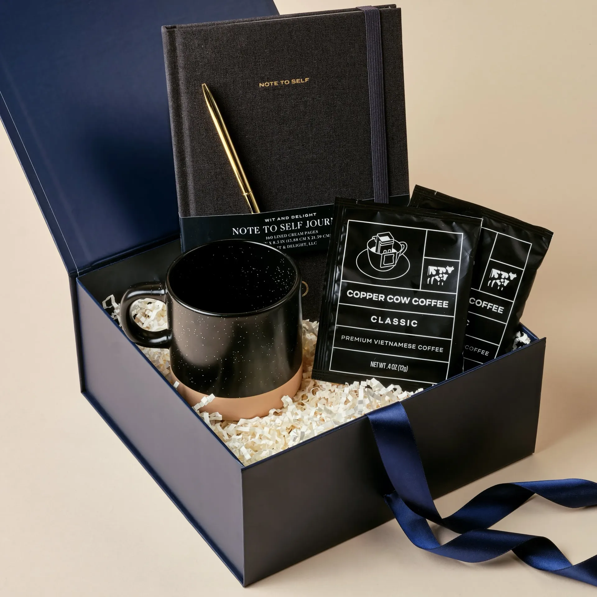 Gift box with a mug, coffee, notebook, pen, and ribbon. Black, gold, and beige tones.