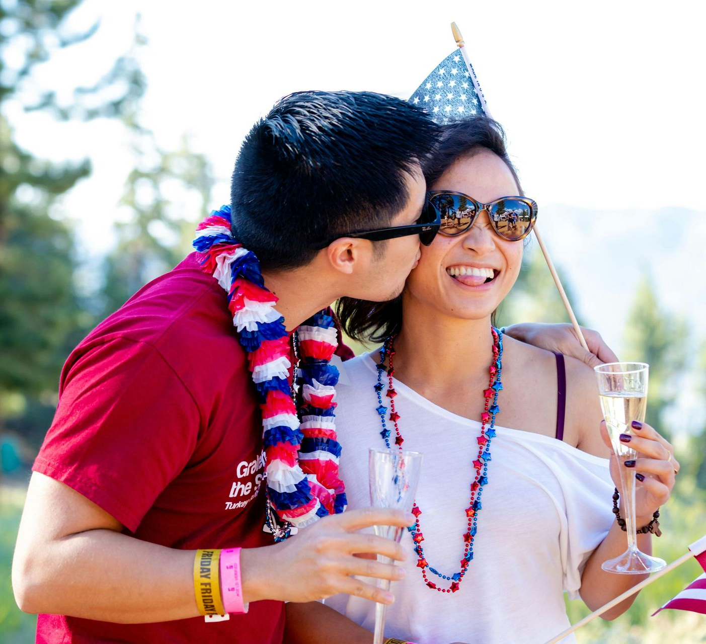 Man kissing woman on the cheek, celebrating outdoors. Both wear patriotic attire, holding drinks and flags.