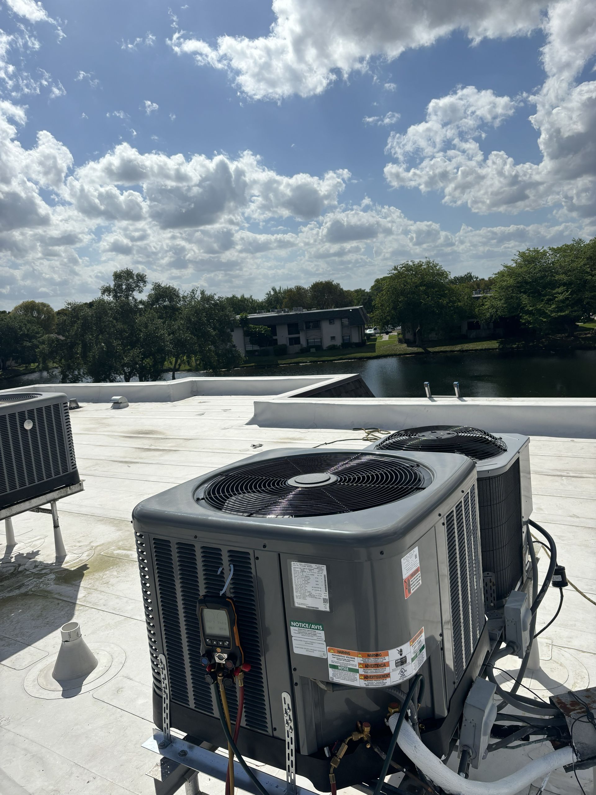 Outdoor HVAC units on a sunny rooftop beside a lake and trees under a cloudy sky