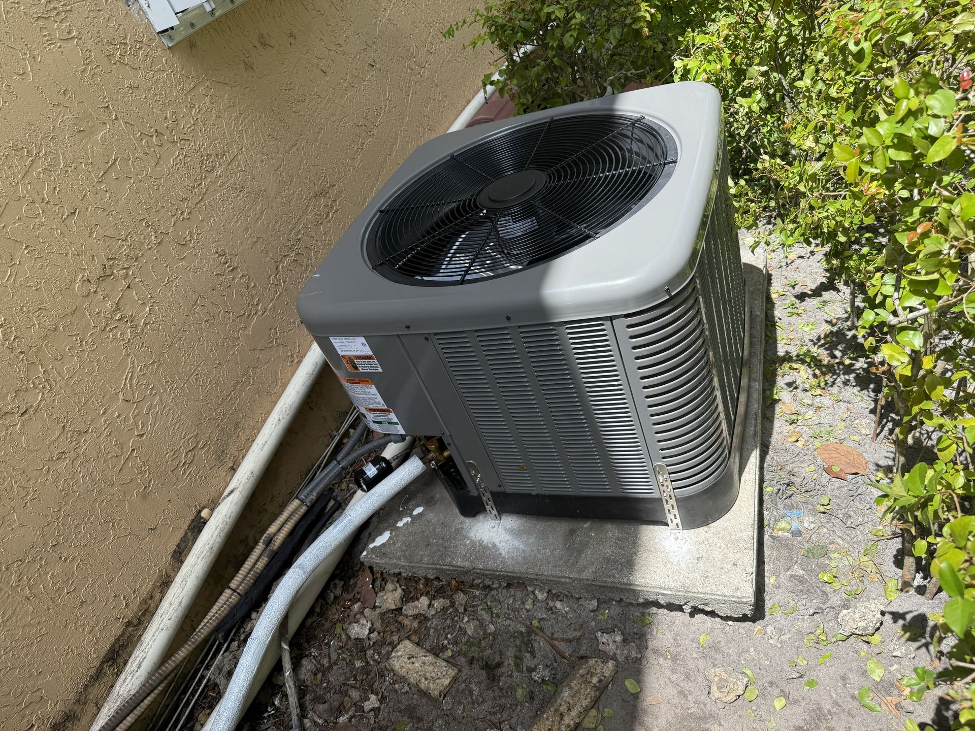 Outdoor air conditioning unit beside a wall, with pipes and cables running along the ground.