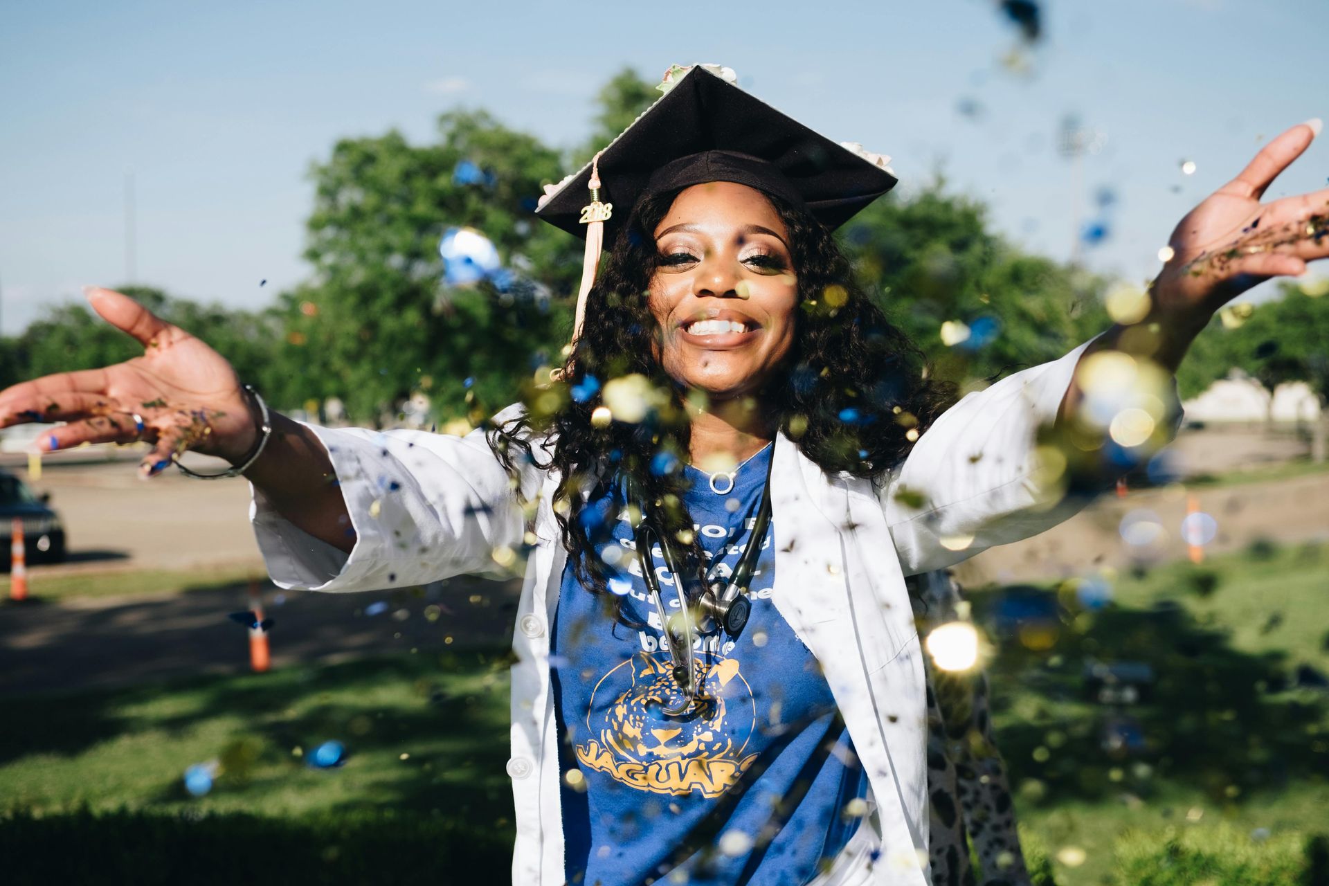 Happy graduate throws confetti in the air, wearing cap and gown outdoors. She is smiling, surrounded by blue and gold confetti.
