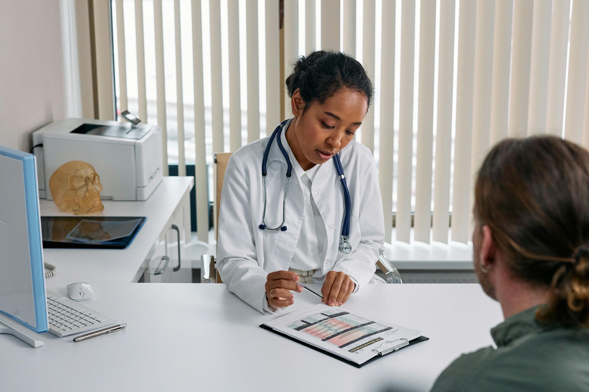 A doctor with a stethoscope examines paperwork at her desk while talking to a male patient.