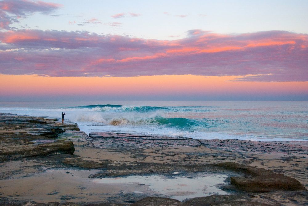 A Person Is Standing on A Rocky Beach Near the Ocean at Sunset — Leisure Coast Powder Coaters in  Ulladulla, NSW