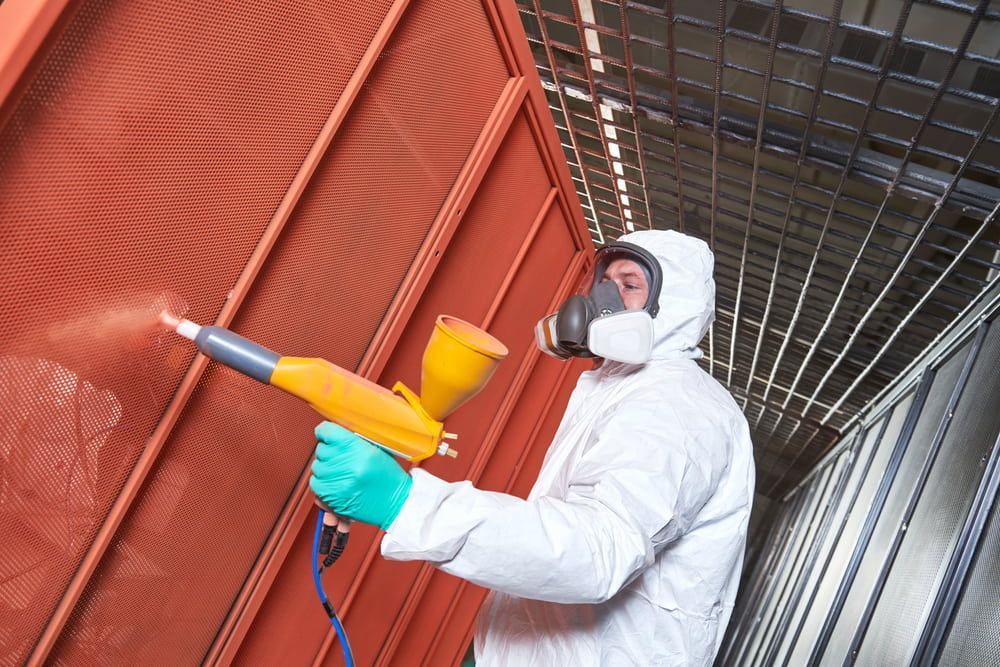 A Man in A Protective Suit Is Spraying Paint on A Wall — Leisure Coast Powder Coaters in Unanderra, NSW
