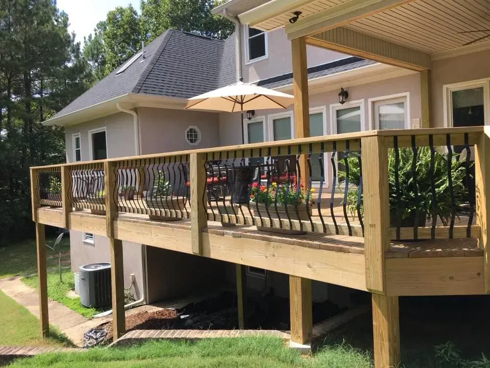 A wooden deck attached to a two-story house with black railing, potted plants, and an open umbrella.