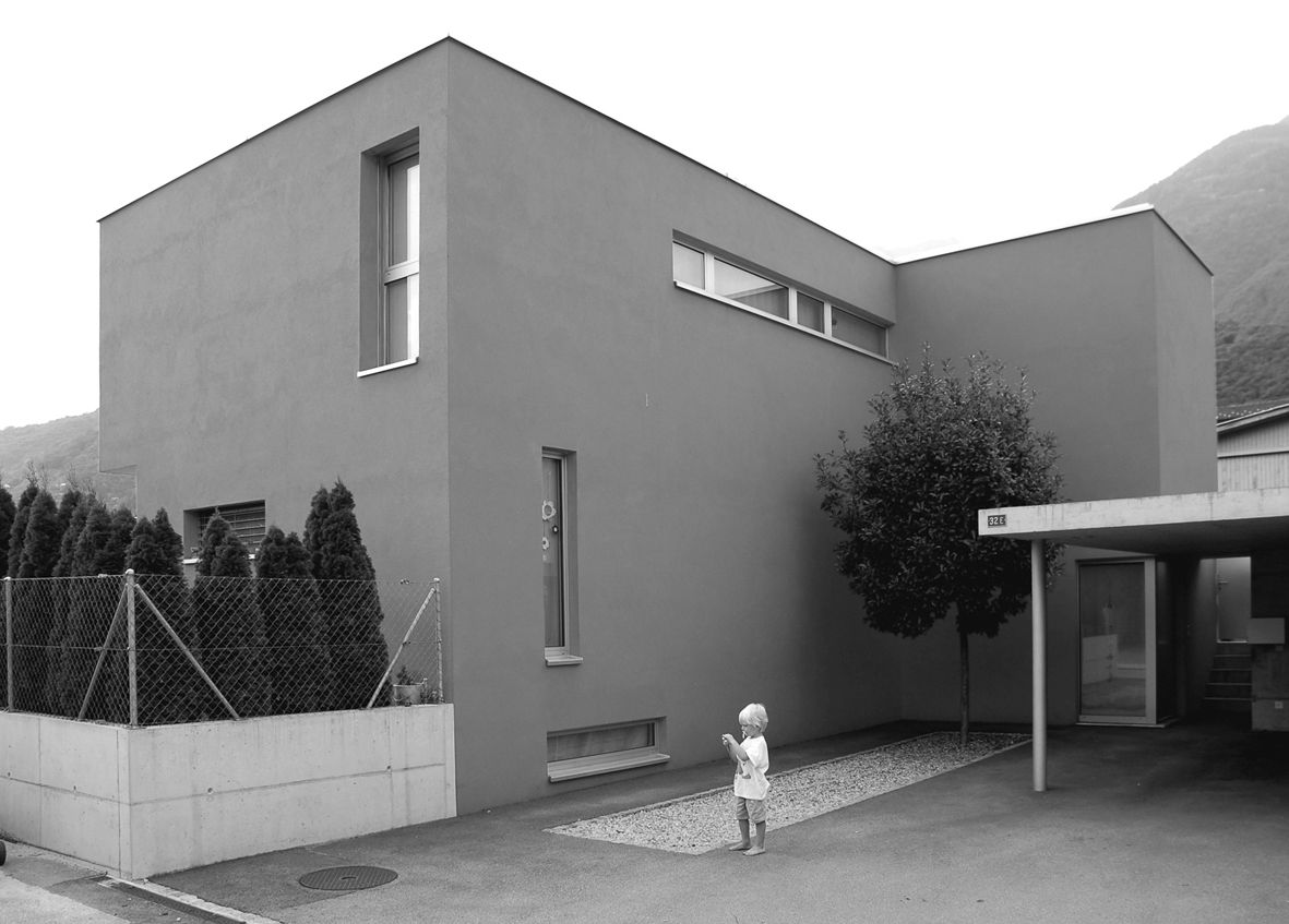 Moderna casa grigia con forme rettangolari, numerose finestre, un bambino piccolo in piedi di fronte, posto auto coperto.