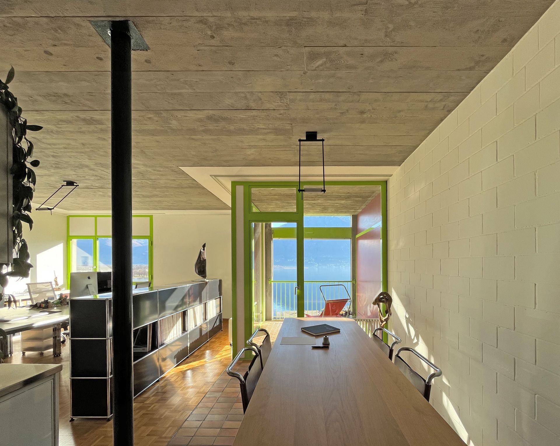 Interni moderni di un ufficio, tavolo e infissi verde brillante che si affacciano su una vista panoramica di lago e montagne.