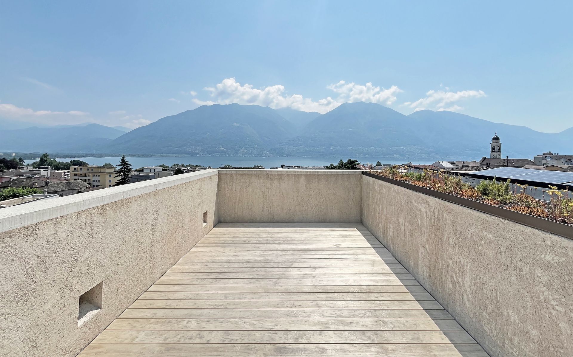 Tetto con pareti strutturate e vista su un lago e montagne sotto un cielo azzurro, paesaggio urbano con un campanile.