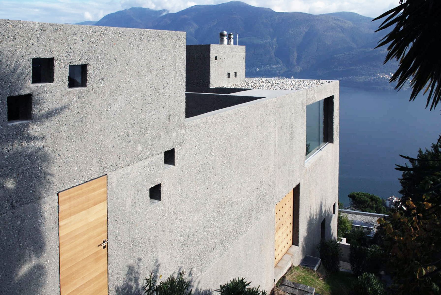 Casa in cemento con finestre quadrate, porte in legno e vista sul lago di montagna.
