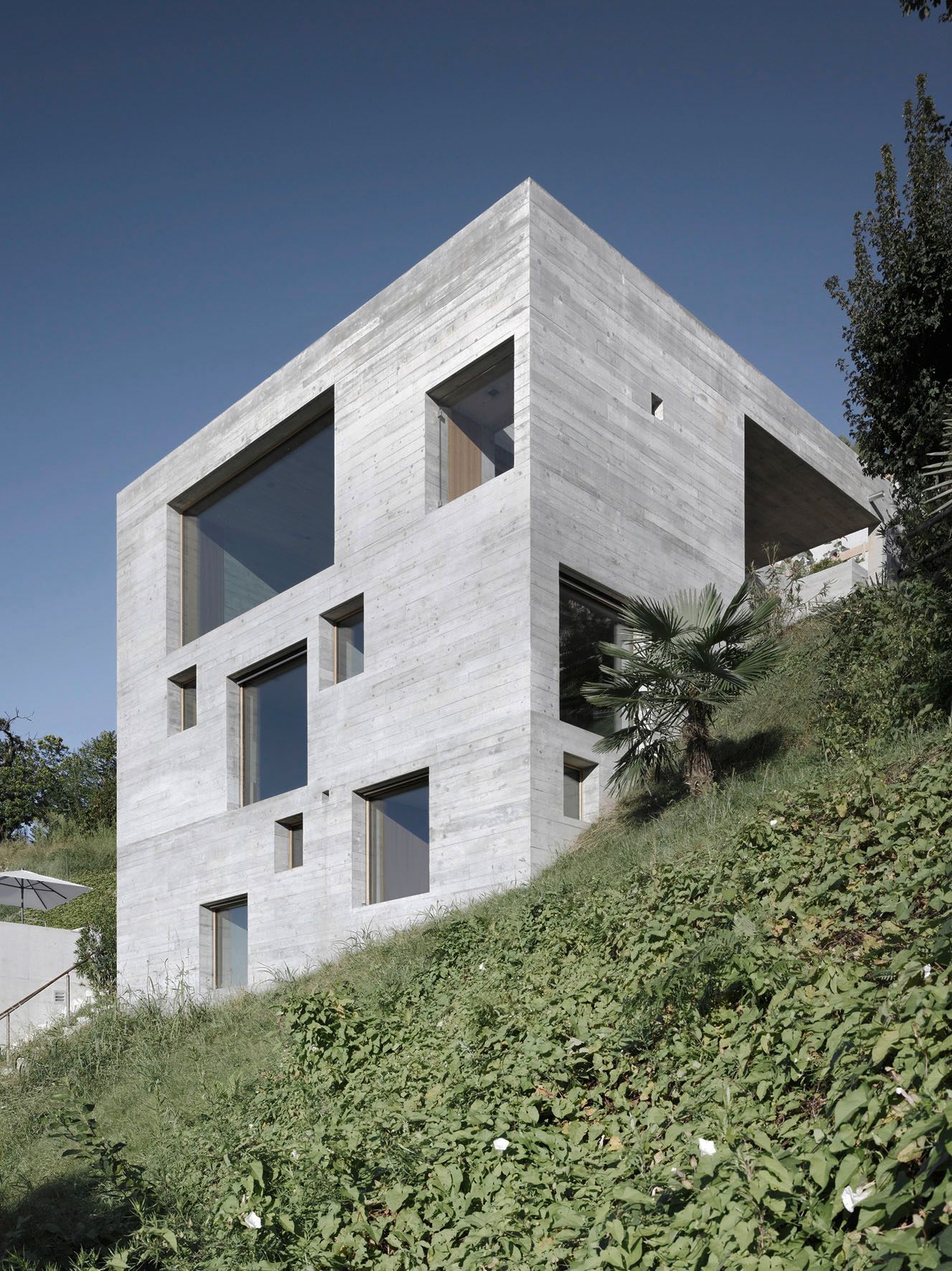 Casa cubica in cemento con diverse finestre rettangolari su un pendio ricoperto di vegetazione verde.