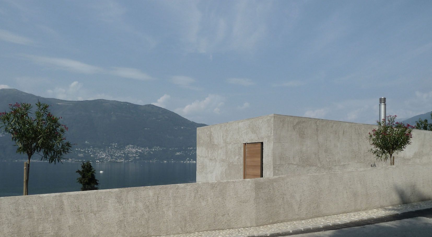 Edificio in cemento con una porta in legno, con vista su un lago e sulle montagne sotto un cielo azzurro.